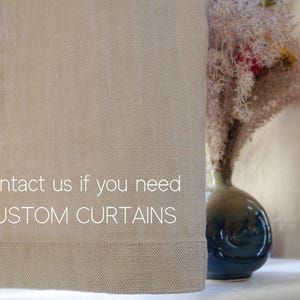 May include: A close-up of a pair of beige linen curtains with the text "Contact us if you need CUSTOM CURTAINS" printed on them. The curtains are hanging in a window with a vase of dried flowers in the background.