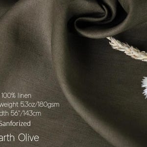 K&ouml;nnte beinhalten: Erdolivenfarbener Leinenstoff, 143 cm breit, mit Text, der darauf hinweist, dass er aus 100 % Leinen, mittlerem Gewicht und sanforisiert ist. Der Stoff ist drapiert, mit einem Zweig aus getrocknetem Weizen und wei&szlig;en Blumen in der Ecke.