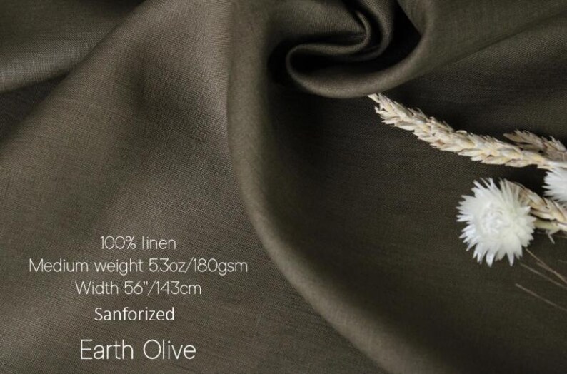 K&ouml;nnte beinhalten: Erdolivenfarbener Leinenstoff, 143 cm breit, mit Text, der darauf hinweist, dass er aus 100 % Leinen, mittlerem Gewicht und sanforisiert ist. Der Stoff ist drapiert, mit einem Zweig aus getrocknetem Weizen und wei&szlig;en Blumen in der Ecke.