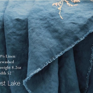 Pu&ograve; includere: Un pezzo di tessuto di lino blu ottanio profondo con una trama visibile e bordi sfilacciati. Il testo sul tessuto recita: 100% Linen, Prewashed, Heavyweight 8.2oz, Width 52'', Forest Lake. Il materiale &egrave; drappeggiato in morbide pieghe.