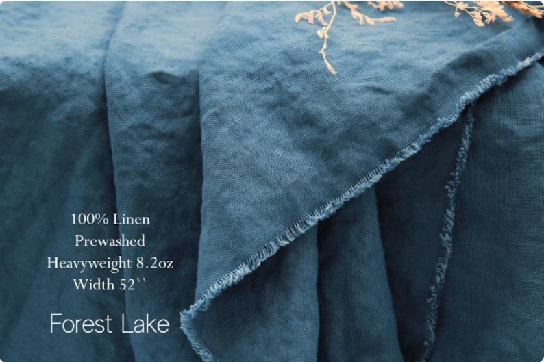 Pu&ograve; includere: Un pezzo di tessuto di lino blu ottanio profondo con una trama visibile e bordi sfilacciati. Il testo sul tessuto recita: 100% Linen, Prewashed, Heavyweight 8.2oz, Width 52'', Forest Lake. Il materiale &egrave; drappeggiato in morbide pieghe.