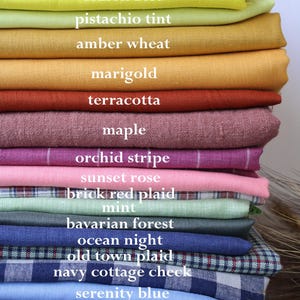 May include: A stack of folded fabric swatches in various colors and patterns, labeled with text including autumn check plaid, lemon zest, pistachio tint, amber wheat, marigold, terracotta, maple, orchid stripe, sunset rose, brick red plaid, mint, bavarian forest, ocean night, old town plaid, navy cottage check, serenity blue, stormy teal, and twilight denim.