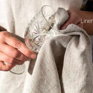 May include: A person is drying a clear glass with a linen towel. The towel is beige and has a rough texture. The glass has a decorative pattern.