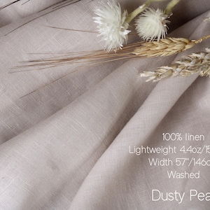 May include: A piece of dusty pearl colored 100% linen fabric with a lightweight 4.4oz weight and a 57 inch width. The fabric is washed and features a visible woven texture, accompanied by dried white flowers and wheat stalks resting on the folds.
