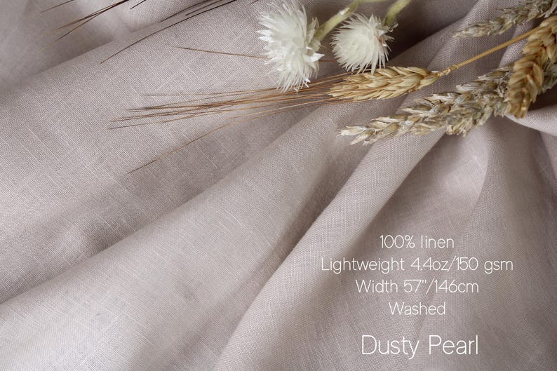 May include: A piece of dusty pearl colored 100% linen fabric with a lightweight 4.4oz weight and a 57 inch width. The fabric is washed and features a visible woven texture, accompanied by dried white flowers and wheat stalks resting on the folds.