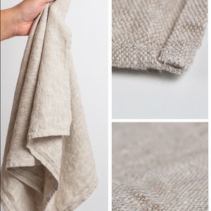 May include: A beige linen tea towel with a textured weave. The towel is folded and held in a hand. The towel has a hemmed edge.