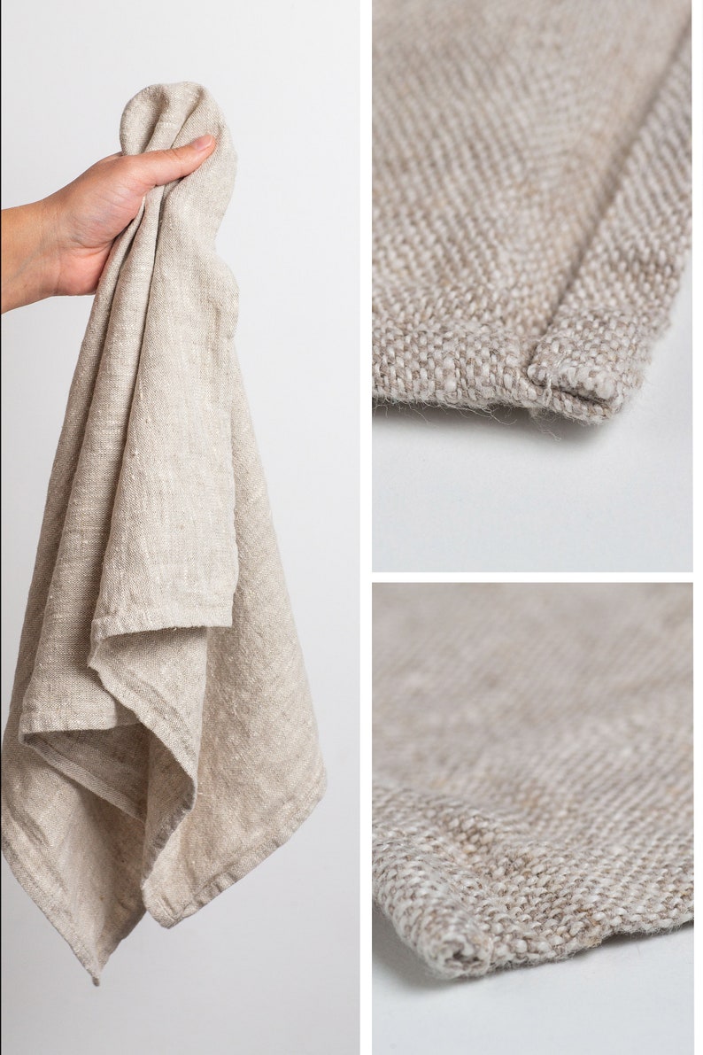 May include: A beige linen tea towel with a textured weave. The towel is folded and held in a hand. The towel has a hemmed edge.