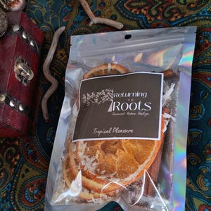 May include: A clear, iridescent package containing dried orange slices, cinnamon sticks, and shredded coconut. The package has a brown label with the text "Returning to Roots" and "Tropical Pleasure". The background is a colorful paisley fabric.
