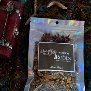 May include: A clear, holographic bag filled with dried herbs and star anise. A black label reads "Returning to Roots" and "Moon Phases". The bag is on a colorful patterned surface, with a red box and decorative spoon in the background.