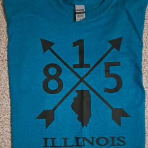 May include: A teal blue t-shirt with a black graphic design. The graphic features the numbers 815, two arrows pointing in opposite directions, and the outline of the state of Illinois. The text "ILLINOIS" is printed below the graphic.