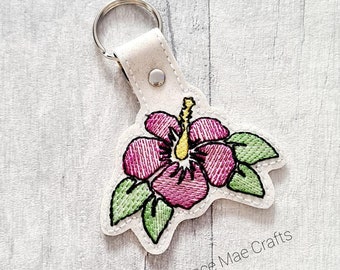 May include: A white leather keychain with a pink hibiscus flower embroidered on it. The flower has green leaves and a yellow centre. The keychain has a silver key ring and the text "Florence Mae Crafts" is printed on the bottom.