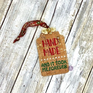 Puede incluir: Una etiqueta de regalo de corcho con un mensaje bordado en rojo y verde que dice "HAND MADE AND IT TOOK ME FOREVER". La etiqueta está atada con una cinta roja y blanca.