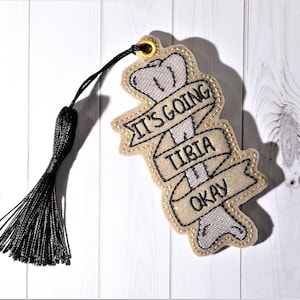 May include: A beige fabric bookmark with a bone design and the text "It's going tibia okay". The bookmark has a black tassel.