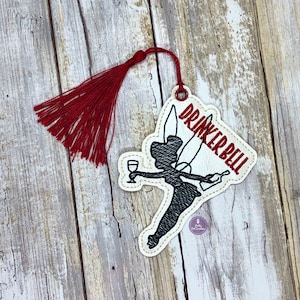 May include: A white fabric ornament with a black silhouette of a fairy holding a wine glass and a bottle. The text "DRINKERBELL" is written in red on the ornament. The ornament has a red tassel.
