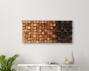 Op de afbeelding: Een houten wandkunstwerk met een verloop van bruine en zwarte vierkanten. De kunst hangt aan een witte muur boven een wit kastje met drie deuren.