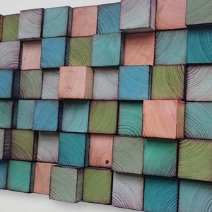 Op de afbeelding: Een wandkunstwerk bestaande uit talrijke houten blokken, elk beschilderd in een andere kleur. De kleuren omvatten tinten blauw, groen en perzik. De blokken zijn in een rasterpatroon gerangschikt, waardoor een textuur en driedimensionaal effect ontstaat. De houtnerf is zichtbaar.