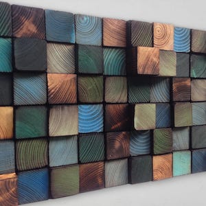 Op de afbeelding: Een wandkunstwerk gemaakt van houten blokken in verschillende tinten blauw, groen en bruin. De blokken zijn in een willekeurig patroon gerangschikt, waardoor een uniek en modern ontwerp ontstaat.