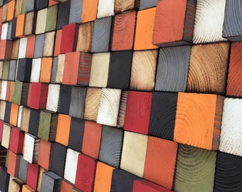 May include: A wall art piece composed of various sized wooden blocks. The blocks are painted in a variety of colors, including red, orange, gray, black, white, and green. The blocks are arranged in a staggered pattern, creating a textured surface.