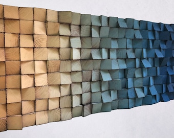 May include: A wall art piece made of wood blocks, painted in a gradient of colors from light brown to dark blue. The blocks are arranged in a staggered pattern, creating a textured surface.