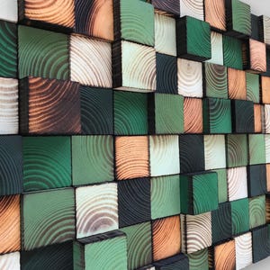 Op de afbeelding: Een wandkunstwerk bestaande uit houten blokken in verschillende kleuren: groen, bruin, zwart en natuurlijke tinten. De blokken zijn in een rasterpatroon gerangschikt, wat een textuur en driedimensionaal effect creëert. De houtnerf is zichtbaar.