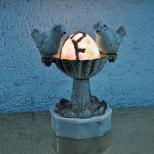 May include: A decorative lamp featuring two white birds perched on a weathered, ornate bowl. The bowl holds a glowing, egg-shaped light with a dark, patterned design. The lamp sits on a white, octagonal base.