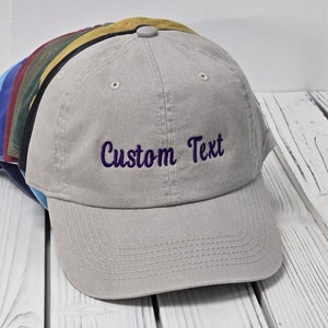 May include: A light gray baseball cap with a purple embroidered text that says "Custom Text". The cap is sitting on a white wooden surface.
