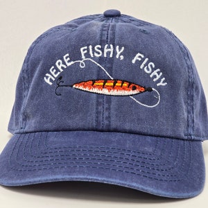 May include: A blue denim baseball cap with an embroidered fishing lure and the text "Here Fishy, Fishy".