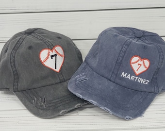 May include: Two distressed denim baseball caps with a red heart and baseball graphic. One cap has the number "7" embroidered on the heart, the other has the name "MARTINEZ" embroidered below the heart.