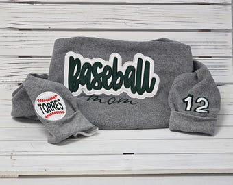 May include: Gray sweatshirt with a green "Baseball Mom" applique and a gray baseball cap with a white "12" on it. A gray beanie with a white baseball and the word "TORRES" is also included.