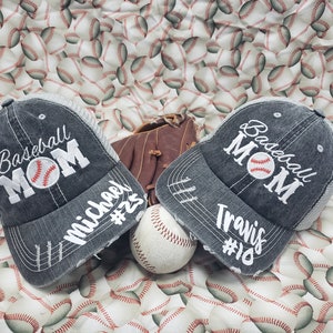 May include: Two grey baseball caps with the text "Baseball Mom" and a baseball graphic. The caps have white text that says "Michael #23" and "Travis #18". A brown baseball glove and a white baseball are in the centre of the image.