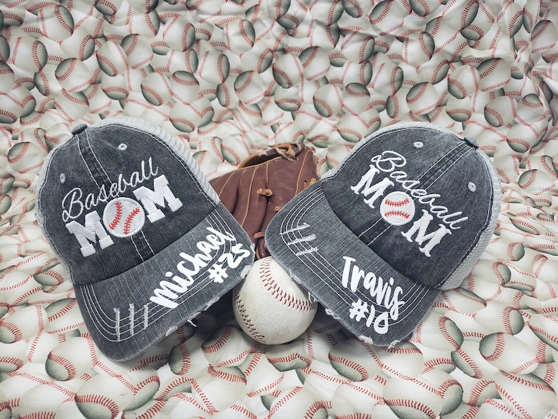 May include: Two grey baseball caps with the text "Baseball Mom" and a baseball graphic. The caps have white text that says "Michael #23" and "Travis #18". A brown baseball glove and a white baseball are in the centre of the image.