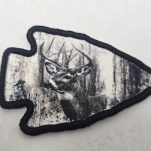 May include: Arrowhead-shaped patch featuring a detailed grayscale image of a deer in a forest setting. The deer has prominent antlers, and the background shows a dense woodland. The patch is outlined with a black border.