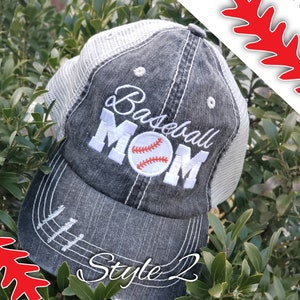 May include: A black and white baseball cap with a mesh back. The cap has the words "Baseball Mom" embroidered on the front in white with a white baseball with red stitching in the centre of the word "Mom". The cap has a distressed look with white stitching around the brim and a white stitching line down the centre of the crown. The cap is sitting on a bed of green leaves. The words "Style 2" are written in white on the brim of the cap.