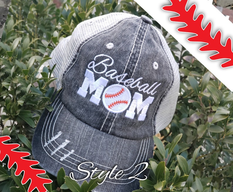 May include: A black and white baseball cap with a mesh back. The cap has the words "Baseball Mom" embroidered on the front in white with a white baseball with red stitching in the centre of the word "Mom". The cap has a distressed look with white stitching around the brim and a white stitching line down the centre of the crown. The cap is sitting on a bed of green leaves. The words "Style 2" are written in white on the brim of the cap.