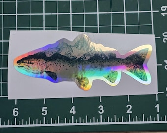 May include: A holographic sticker of a trout with a mountain range in the background. The sticker has a rainbow iridescent finish.