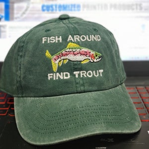 May include: A dark green baseball cap with the embroidered text "FISH AROUND FIND TROUT" and a colorful trout illustration. The hat is made of a soft, worn material, perfect for fishing enthusiasts.