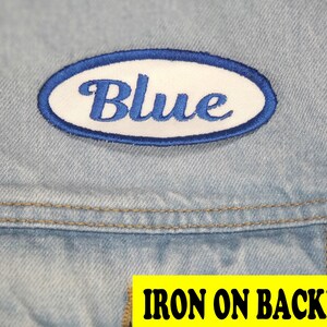 May include: A blue embroidered patch with the word "Blue" in script lettering, set within a white oval with a blue border. The patch is on a light blue denim fabric. The text "IRON ON BACKING" is in a yellow rectangle.