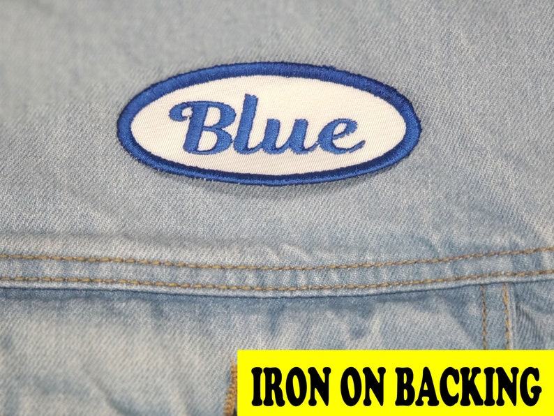 May include: A blue embroidered patch with the word "Blue" in script lettering, set within a white oval with a blue border. The patch is on a light blue denim fabric. The text "IRON ON BACKING" is in a yellow rectangle.