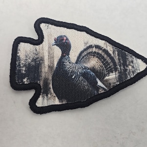 May include: Arrowhead-shaped patch featuring a detailed image of a dark-colored bird with a fanned tail, set against a blurred forest backdrop. The patch has a black border, and the bird appears to be a turkey or similar fowl.
