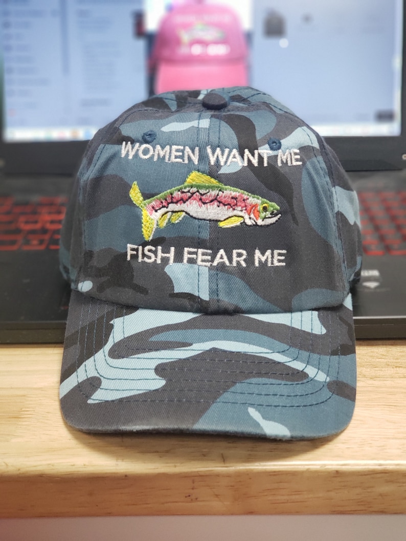 May include: A blue camouflage baseball cap with the text "Women Want Me" and "Fish Fear Me" embroidered on the front. A colorful fish is embroidered between the text.