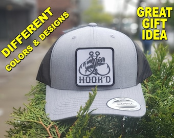 May include: Gray and black trucker hat with a white square patch featuring a black and white illustration of a fish with two hooks. The patch says "HOOK'D".