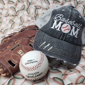 May include: A grey baseball cap with a white mesh back and the text "Baseball Mom" embroidered on the front. A white baseball with red stitching and the text "Super Champion Regulation Official League" is in the foreground. A brown leather baseball glove is in the background.