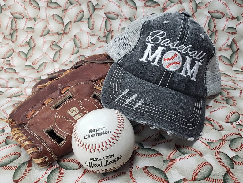 May include: A grey baseball cap with a white mesh back and the text "Baseball Mom" embroidered on the front. A white baseball with red stitching and the text "Super Champion Regulation Official League" is in the foreground. A brown leather baseball glove is in the background.