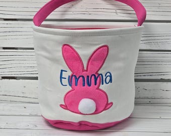 May include: White Easter basket with pink bunny ears and a pink bunny with a white tail. The basket has a pink handle and the name "Emma" is embroidered in blue on the front.