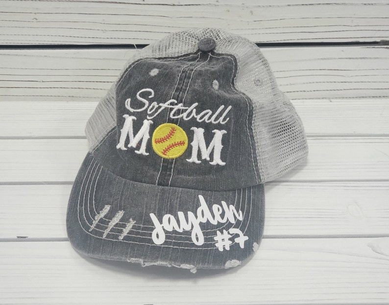 May include: A distressed trucker hat in grey and white with the text "Softball Mom" and a yellow softball with red stitching. The hat also has the name "Jayden" and the number "#7" embroidered on the front.