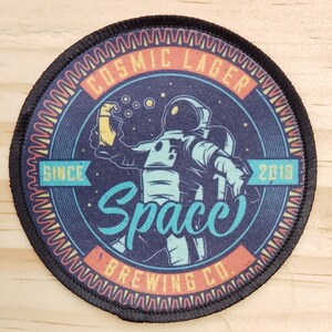 May include: A round, black patch with a colorful design. The design features an astronaut holding a beer with the text "Cosmic Lager" and "Space Brewing Co." around the astronaut. The text "Since 2018" is also included in the design.