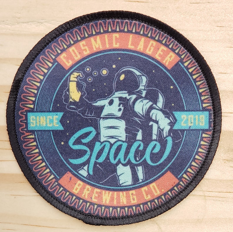 May include: A round, black patch with a colorful design. The design features an astronaut holding a beer with the text "Cosmic Lager" and "Space Brewing Co." around the astronaut. The text "Since 2018" is also included in the design.