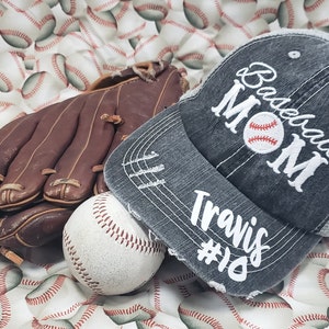 May include: A grey baseball cap with a white distressed design that says "Baseball Mom" with a baseball graphic and "Travis #10" below. The cap is on a white background with a brown baseball glove and a white baseball.