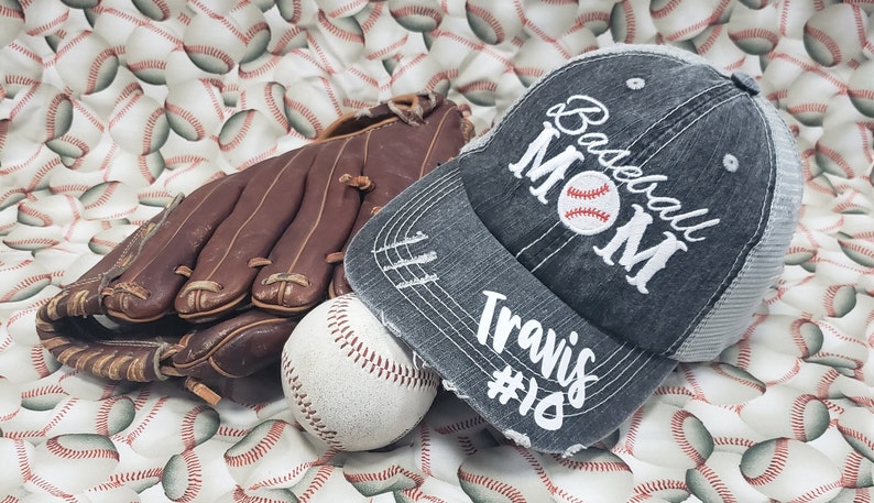 May include: A grey baseball cap with a white distressed design that says "Baseball Mom" with a baseball graphic and "Travis #10" below. The cap is on a white background with a brown baseball glove and a white baseball.