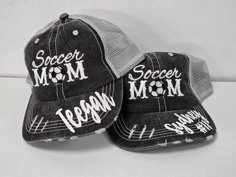 May include: Two black and gray mesh trucker hats with the text "Soccer Mom" embroidered on the front. The hats have a white soccer ball graphic and are personalized with the names "Teegan" and "Sydney".